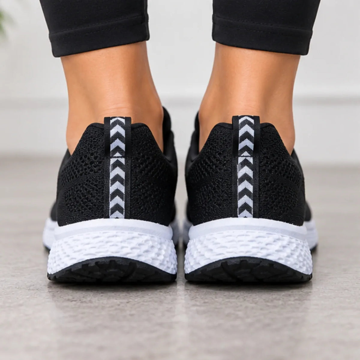 Black knit athletic sneakers with white chunky soles, shown from the back on a person's feet against a light gray indoor floor.