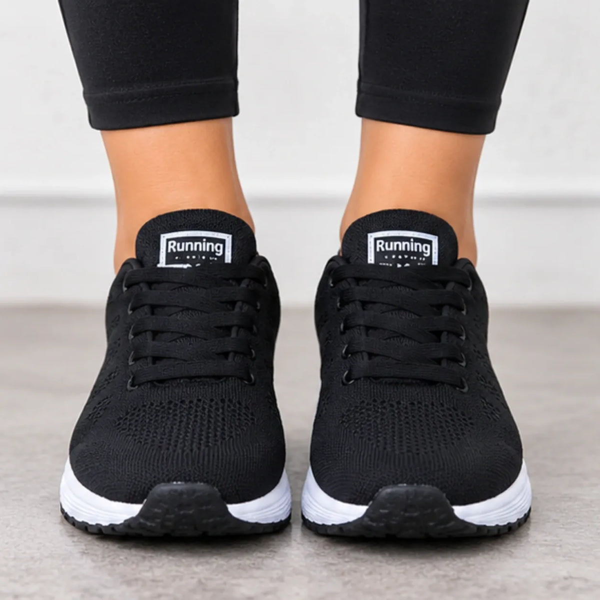 A pair of black athletic sneakers with white midsoles, shown on a person's feet wearing black leggings. The tongue of each shoe displays a white label with the brand name 'Running Machine'.