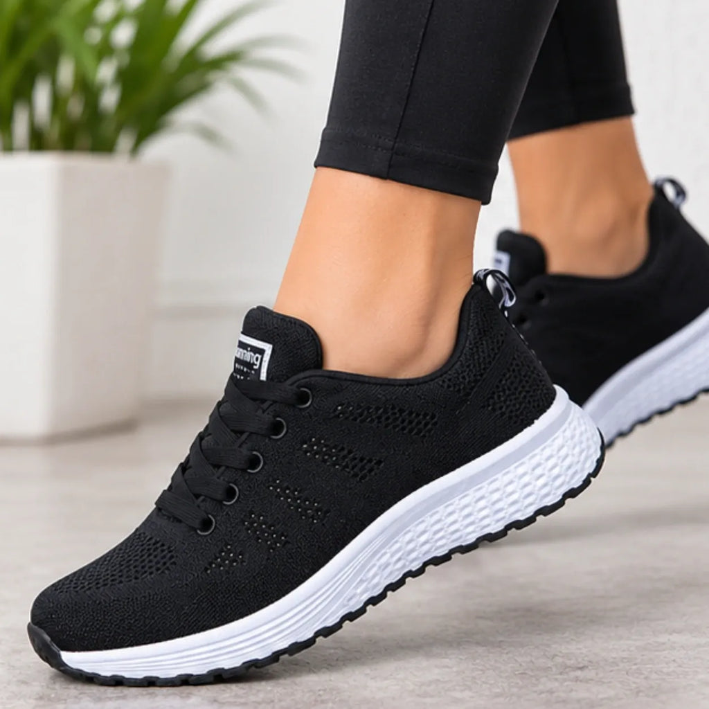 A person wearing black knit athletic sneakers with thick white midsoles, shown from the side on a light indoor floor with a blurred plant in the background.
