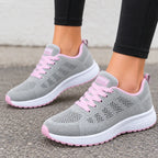 A pair of gray knit athletic sneakers with pink laces, pink trim, and white soles, shown being worn with black leggings on a gray pavement surface.
