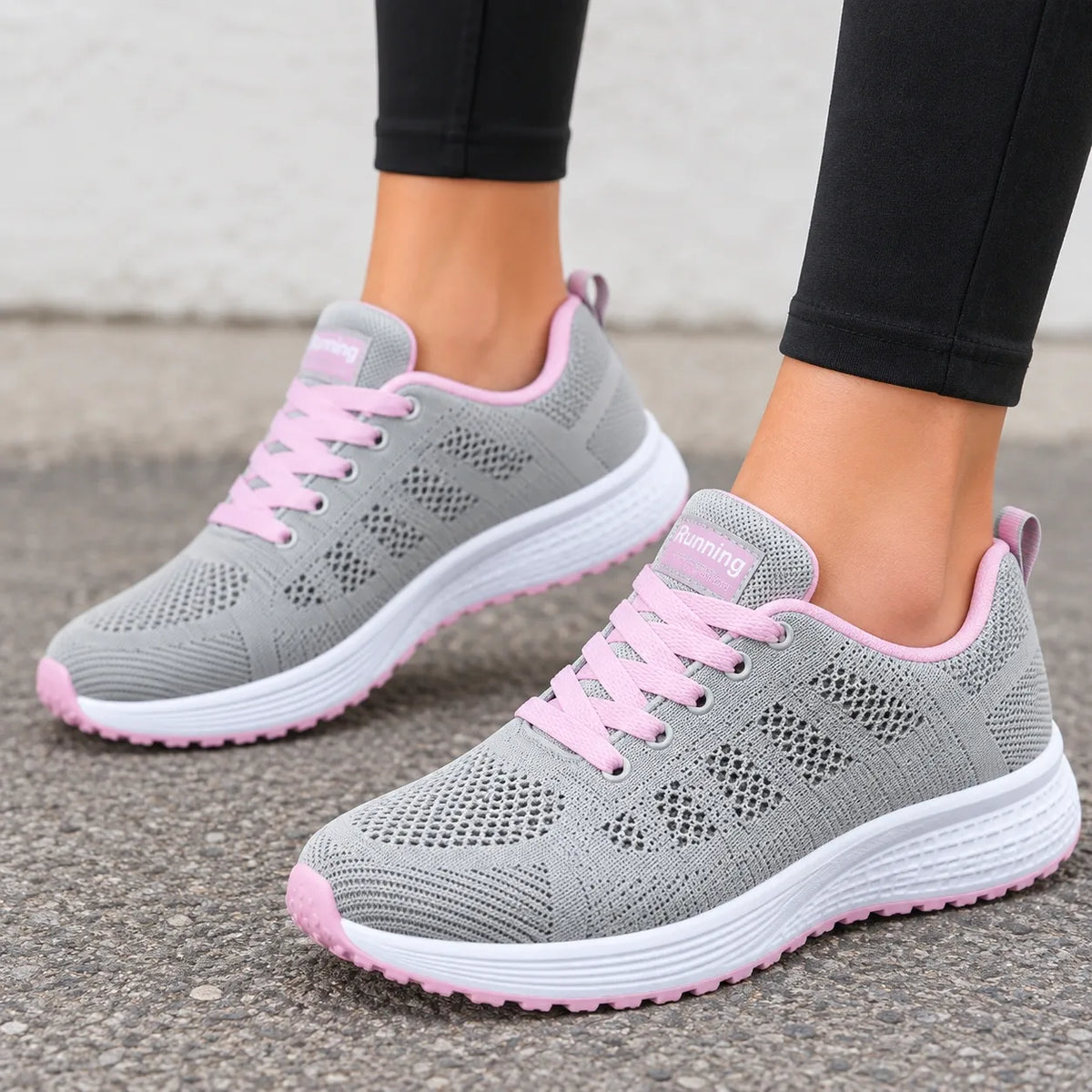 A pair of gray knit athletic sneakers with pink laces, pink trim, and white soles, shown being worn with black leggings on a gray pavement surface.