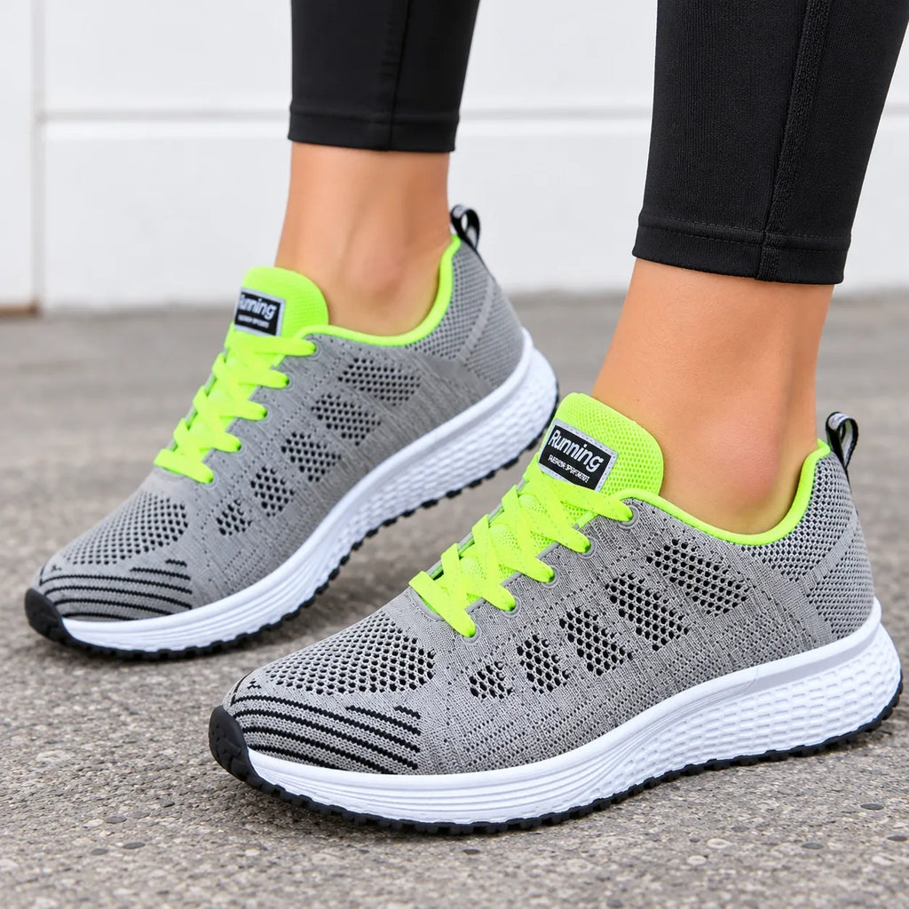 A pair of gray knit athletic sneakers with bright neon green accents and white soles, shown on a person's feet wearing black leggings on a gray pavement surface.