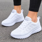 A pair of white athletic sneakers with a breathable mesh upper and thick cushioned soles, shown on a person's feet standing on gray asphalt.