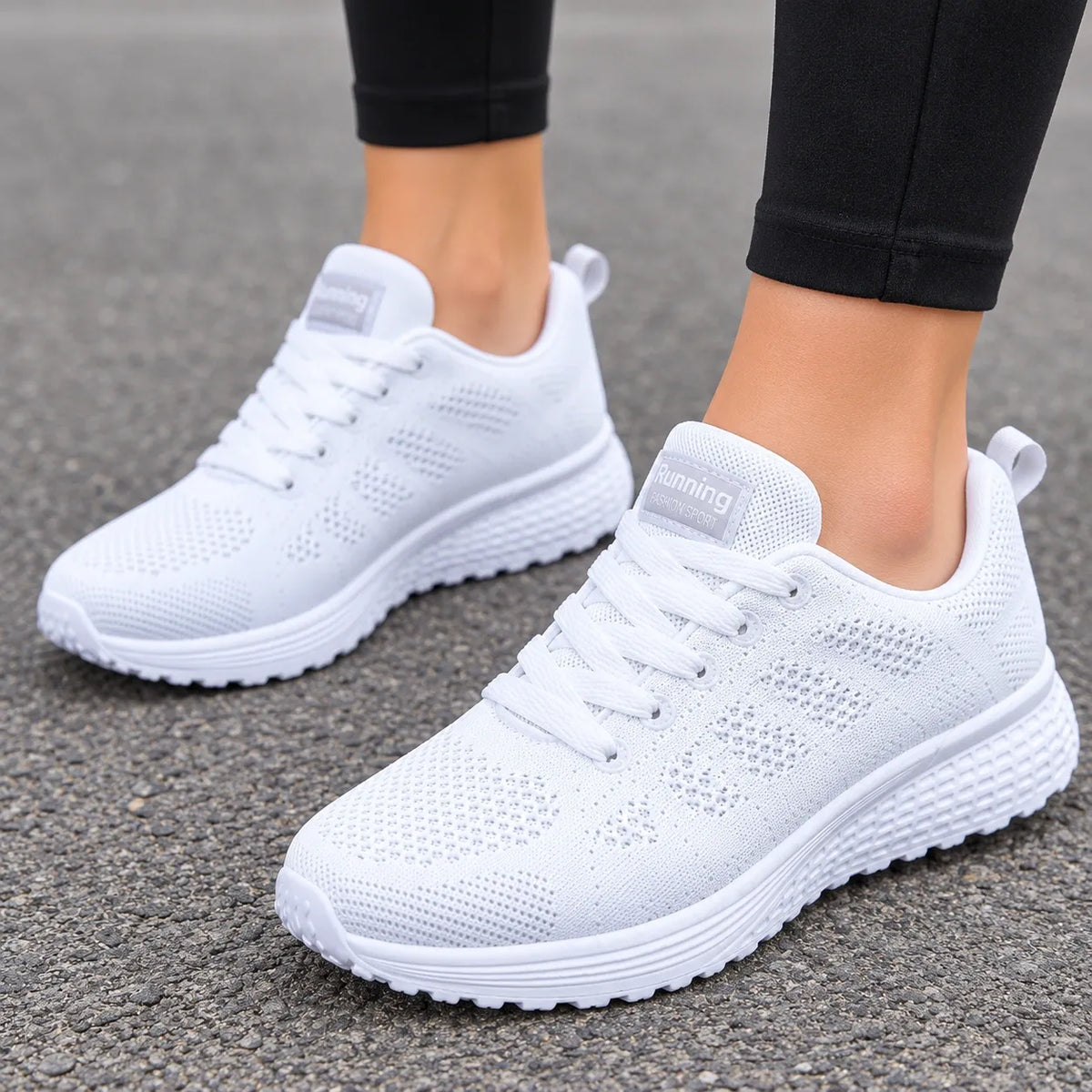 A pair of white athletic sneakers with a breathable mesh upper and thick cushioned soles, shown on a person's feet standing on gray asphalt.