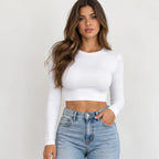 A woman modeling a fitted white long-sleeve crop top paired with light-wash blue jeans against a plain light gray studio background.