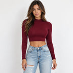 A woman modeling a wine red long-sleeve mock neck crop top paired with light-wash distressed blue jeans against a plain white background.