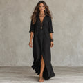A woman modeling a black linen maxi dress with a split front hem and buttoned chest, styled with layered necklaces and bracelets against a textured gray concrete wall.