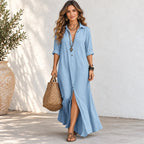 A woman models a light blue linen shirt dress with a high slit, holding a woven straw bag against a white stucco wall background.