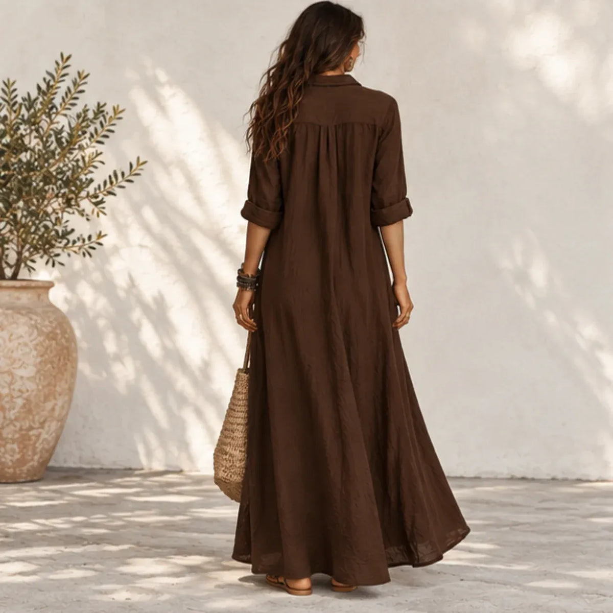 A woman viewed from behind wearing a long brown linen maxi dress with three-quarter sleeves, standing in front of a white stucco wall with leaf shadows. She is holding a woven straw tote bag.