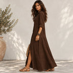 A woman wearing a long brown midi dress with three-quarter sleeves and a side slit, standing in front of a white wall with leaf shadows.