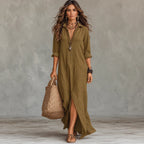 A woman models a long olive green linen shirt dress with rolled sleeves, a button-down front, and a high side slit. She is holding a large woven straw tote bag and wearing layered necklaces, bracelets, and brown sandals, standing against a textured gray studio backdrop.