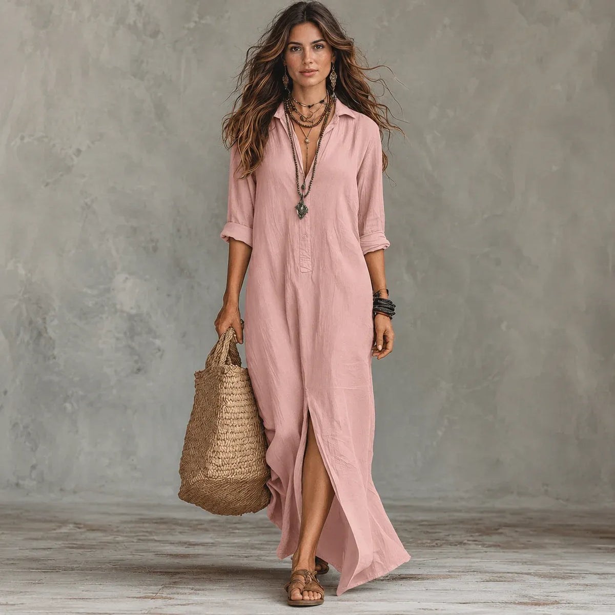 A woman models a dusty pink linen shirt dress with a high slit, holding a woven straw tote bag against a textured gray studio backdrop.