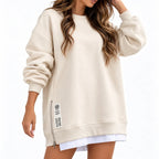 A woman wearing an oversized cream-colored sweatshirt with a side zipper and patch detail, paired with a white shirt peeking out at the hem, set against a plain white background.