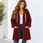 A woman wearing a long burgundy hooded zip-up jacket with white drawstrings, styled over a white tank top and blue jeans, standing against a white wall.