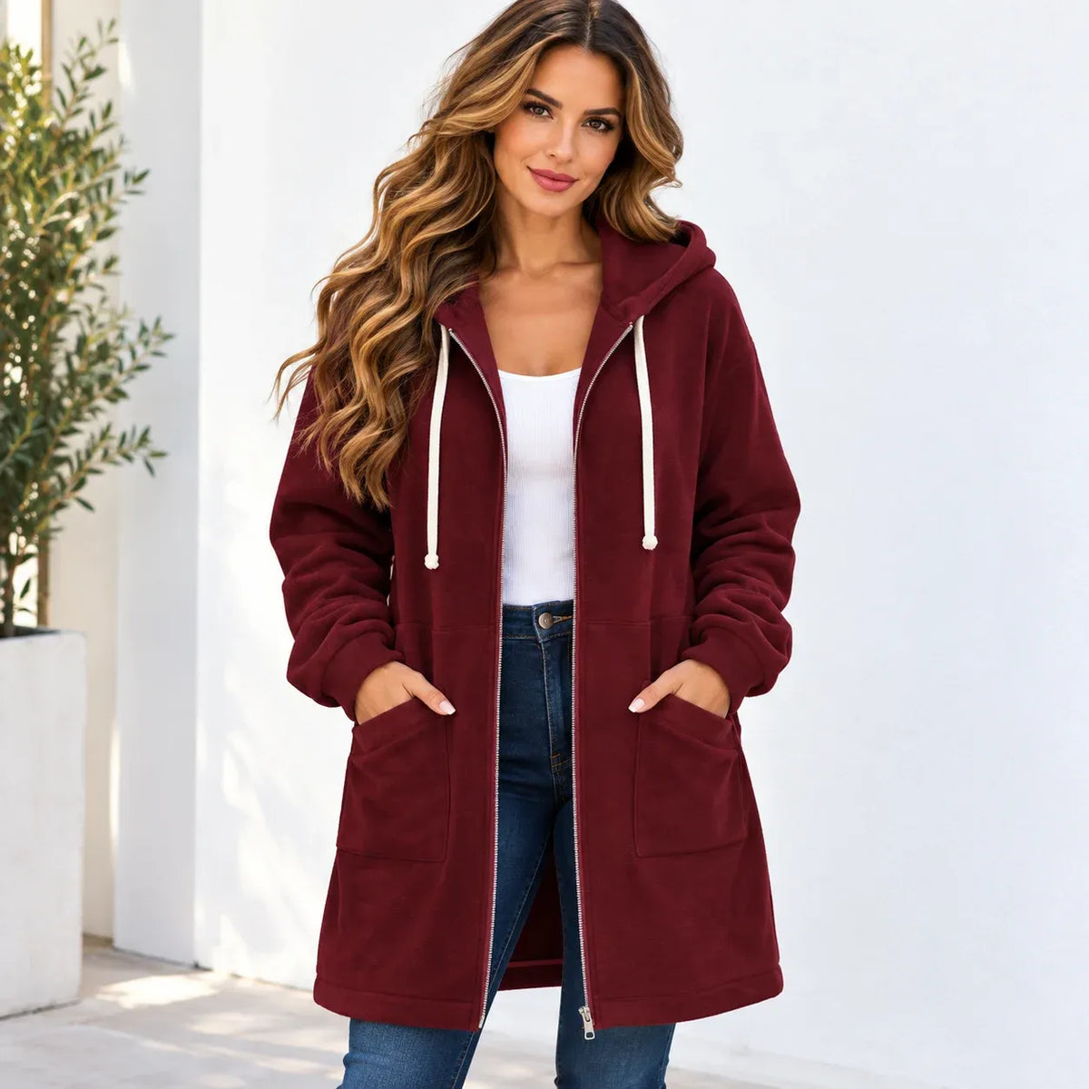 A woman wearing a long burgundy hooded zip-up jacket with white drawstrings, styled over a white tank top and blue jeans, standing against a white wall.
