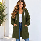 A woman wearing a long olive green zip-up hoodie with a front pocket, styled over a white tank top and blue jeans, standing against a white wall.