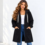 A woman wearing a black mid-length hooded zip-up jacket with white drawstrings, a white tank top, and blue jeans, standing against a white wall.