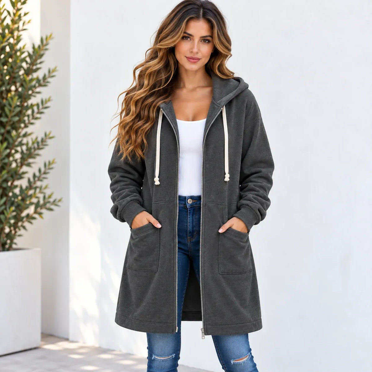 A woman wearing a dark gray hooded zip-up sweatshirt with cream drawstrings, a white tank top, and blue jeans, standing against a white wall with a potted plant on the left.