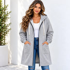 A woman wearing a light gray zip-up hooded sweatshirt with cream drawstrings, a white tank top, and blue jeans, standing against a white wall.