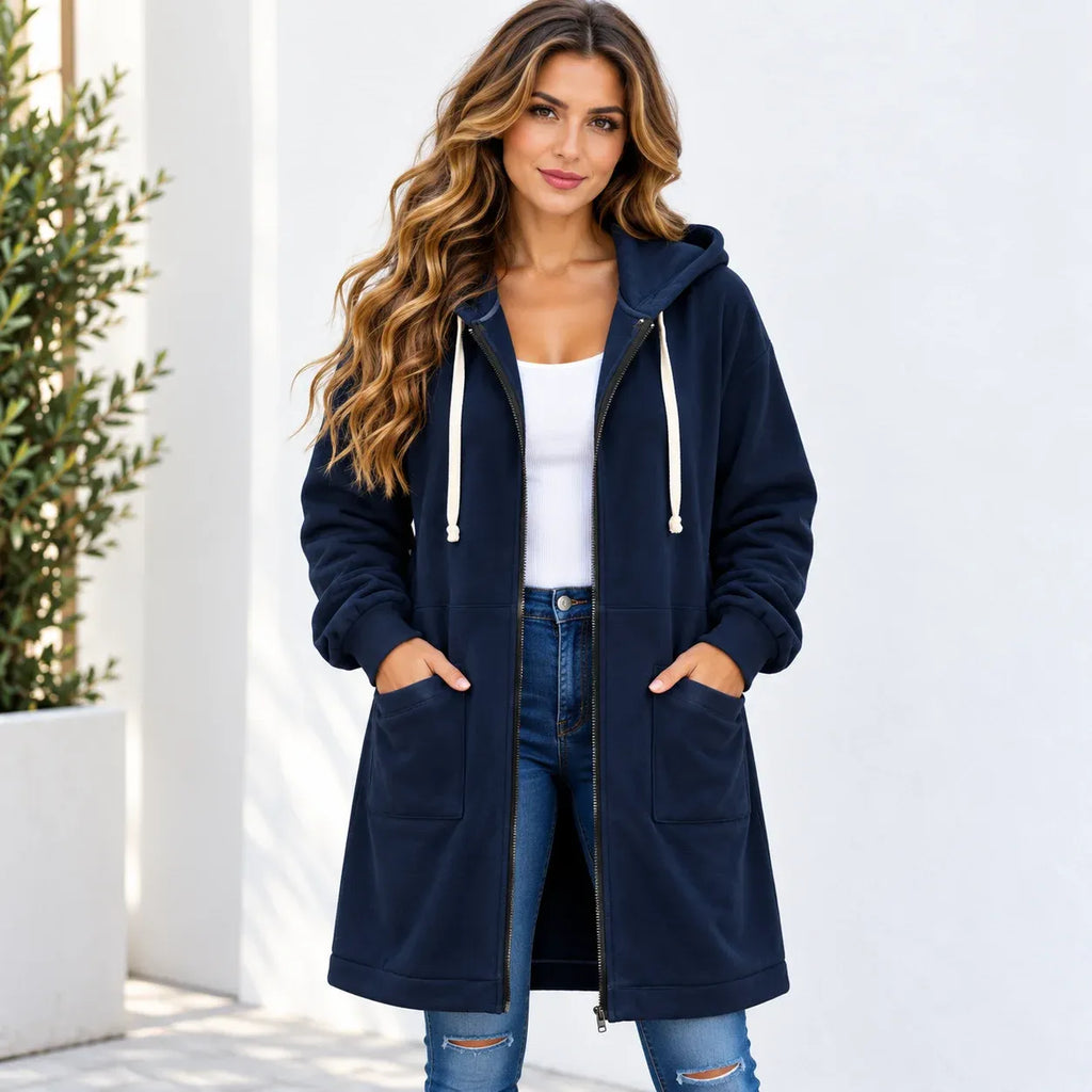 A woman wearing a navy blue hooded zip-up jacket with white drawstrings, a white tank top, and blue jeans, standing against a white wall.