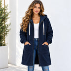 A woman wearing a navy blue hooded zip-up jacket with white drawstrings, a white tank top, and blue jeans, standing against a white wall.