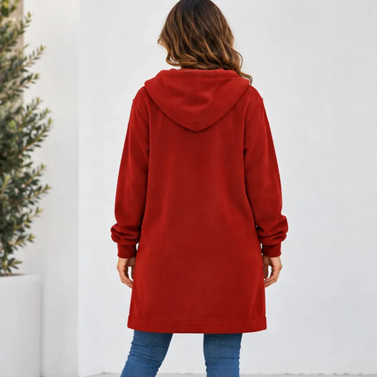 A woman viewed from behind wearing a red hooded sweatshirt and blue jeans, standing against a white wall with a potted plant on the left.