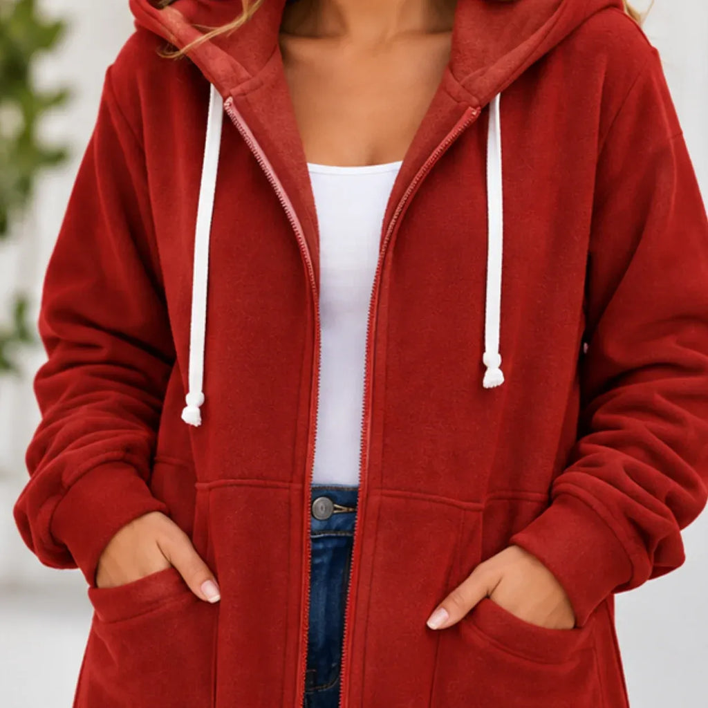 A person wearing a red zip-up hoodie with white drawstrings over a white tank top and blue jeans, shown from the neck down against a softly blurred light background.