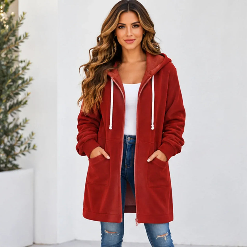 A woman wearing a long red hooded zip-up jacket with white drawstrings, a white tank top, and blue jeans, standing against a white wall.