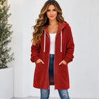 A woman wearing a long red hooded zip-up jacket with white drawstrings, a white tank top, and blue jeans, standing against a white wall.