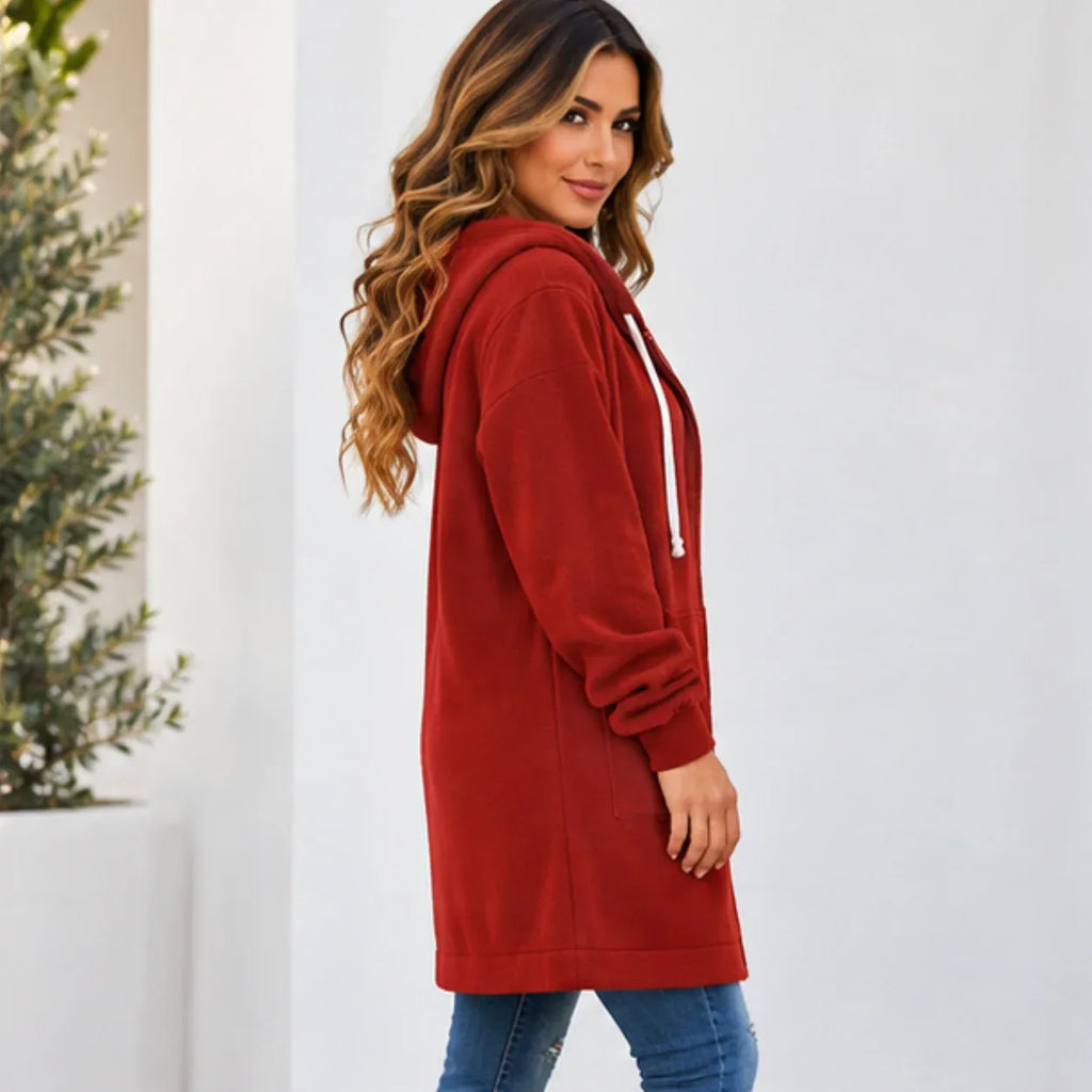 A woman wearing a long red hooded sweatshirt with white drawstrings, paired with blue jeans, standing against a white wall with a potted plant on the left.