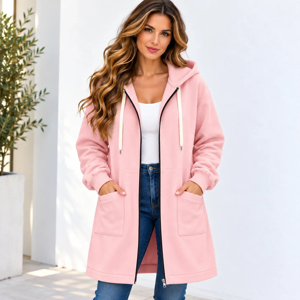 A woman wearing a light pink hooded zip-up jacket with cream drawstrings, a white tank top, and blue jeans, standing against a white wall with a potted plant on the left.