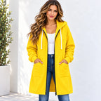 A woman wearing a bright yellow hooded jacket with white drawstrings, a white tank top, and blue jeans, standing against a white wall with a potted plant on the left.