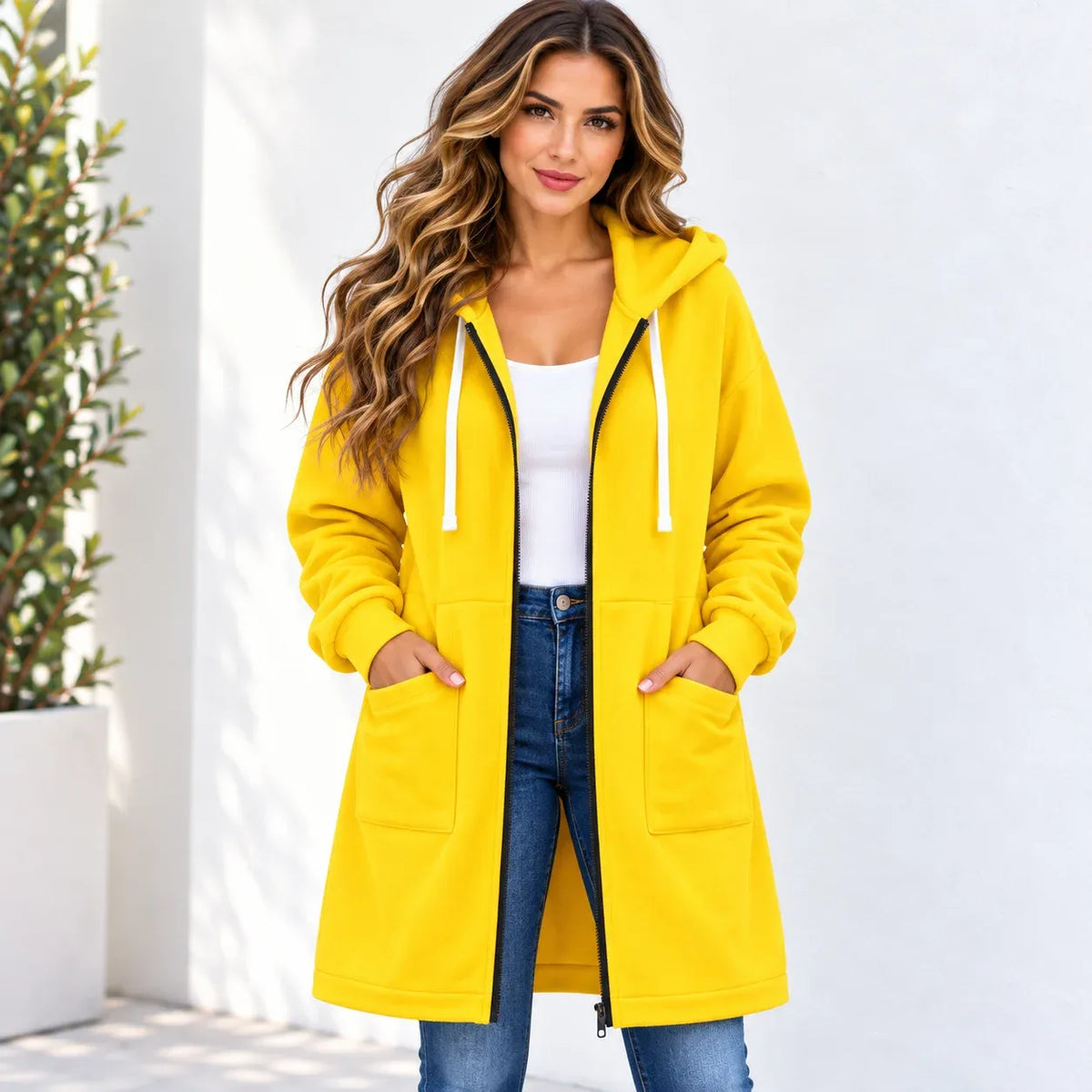 A woman wearing a bright yellow hooded jacket with white drawstrings, a white tank top, and blue jeans, standing against a white wall with a potted plant on the left.