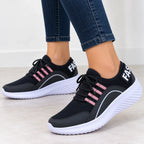 A pair of black mesh sneakers with pink stripe accents and white soles, shown on a model wearing blue jeans against a clean indoor background.