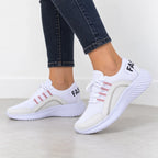 White low-top sneakers with light pink stripe accents and black lettering on the heel, shown on a model wearing dark blue jeans.
