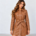 A woman modeling a brown faux-leather trench coat dress with a matching tie belt, photographed against a plain light gray studio background.