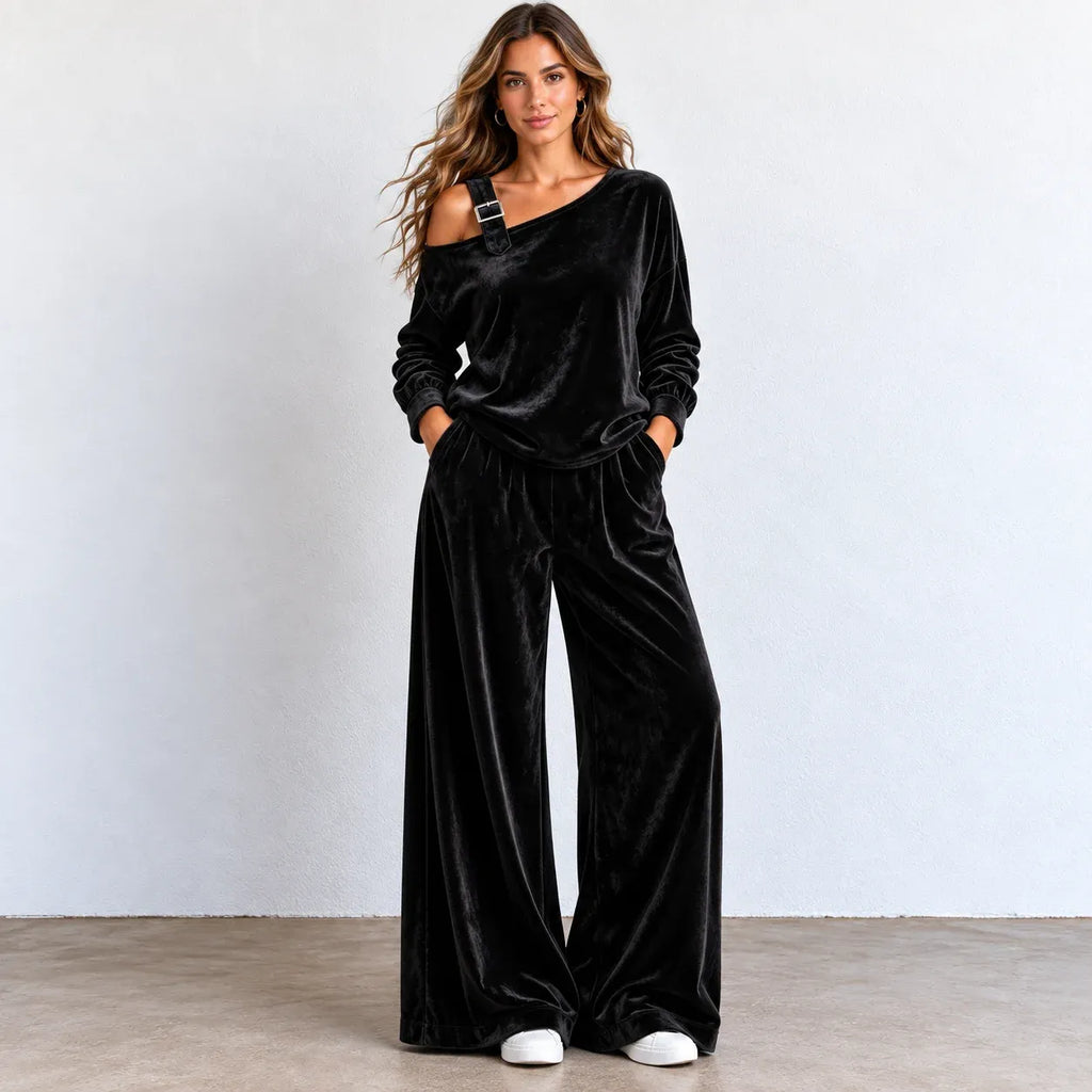 A woman modeling a black velvet two-piece loungewear set consisting of an asymmetrical one-shoulder long-sleeve top and matching wide-leg pants, standing against a light gray wall.