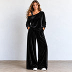 A woman modeling a black velvet two-piece loungewear set consisting of an asymmetrical one-shoulder long-sleeve top and matching wide-leg pants, standing against a light gray wall.