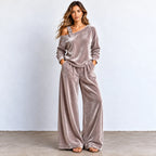 A woman models a matching two-piece lounge set in a dusty mauve velvet fabric. The set includes an asymmetrical one-shoulder long-sleeve top with a decorative strap detail and matching wide-leg pants.