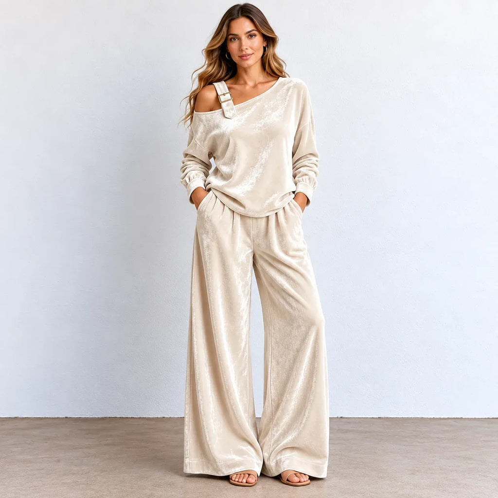 A woman modeling a two-piece lounge set consisting of a long-sleeve off-the-shoulder top and matching wide-leg pants in a champagne-colored velvet fabric, standing against a light gray wall.