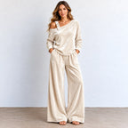 A woman modeling a two-piece lounge set consisting of a long-sleeve off-the-shoulder top and matching wide-leg pants in a champagne-colored velvet fabric, standing against a light gray wall.