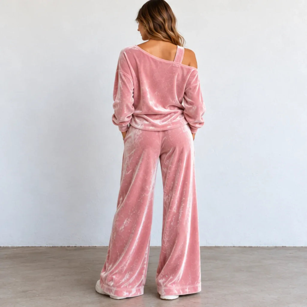 A woman viewed from behind wearing a dusty pink velvet two-piece lounge set with a cold-shoulder long-sleeve top and matching wide-leg pants, standing against a plain light gray wall.