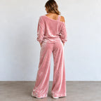 A woman viewed from behind wearing a dusty pink velvet two-piece lounge set with a cold-shoulder long-sleeve top and matching wide-leg pants, standing against a plain light gray wall.