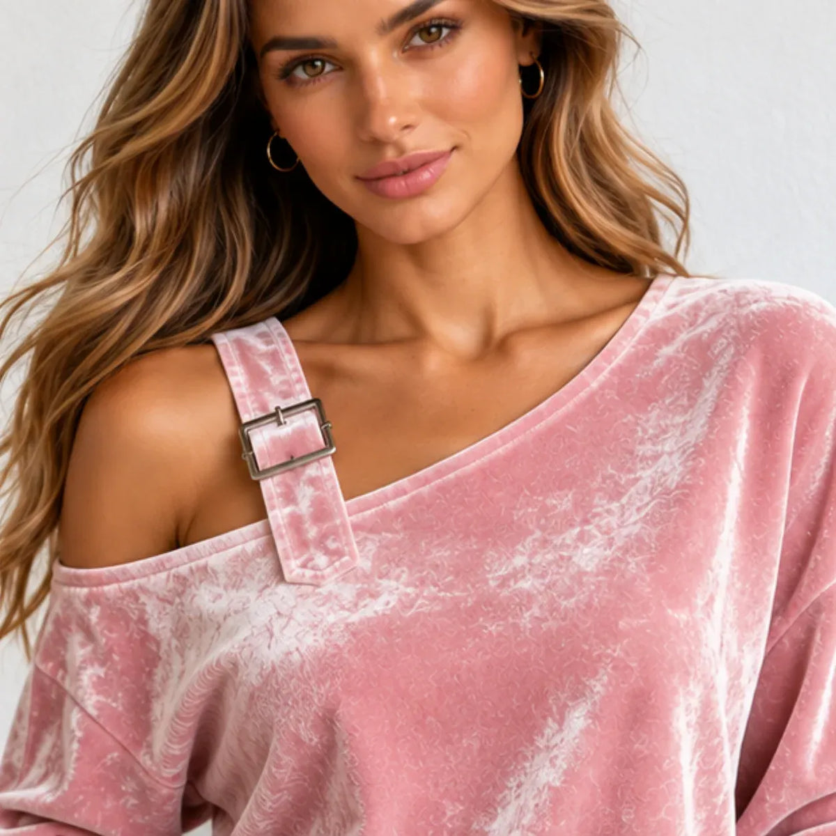 A woman wearing a dusty pink velvet off-the-shoulder top with a wide strap and silver buckle detail, set against a clean light gray background.