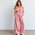 A woman modeling a pink velvet two-piece loungewear set consisting of an asymmetrical one-shoulder long-sleeve top and matching wide-leg pants, standing against a plain light gray wall.