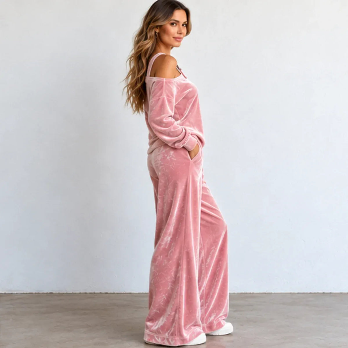 A woman modeling a dusty pink velvet off-the-shoulder jumpsuit with wide legs, standing against a plain light gray wall.