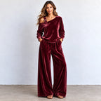 A woman modeling a two-piece velvet loungewear set in deep burgundy. The set includes an asymmetrical one-shoulder long-sleeve top with a decorative strap detail on the shoulder and matching wide-leg pants.