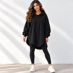 A woman modeling a black oversized pullover hoodie with drawstrings, paired with black leggings and white sneakers, standing against a white textured wall with soft diagonal shadows.