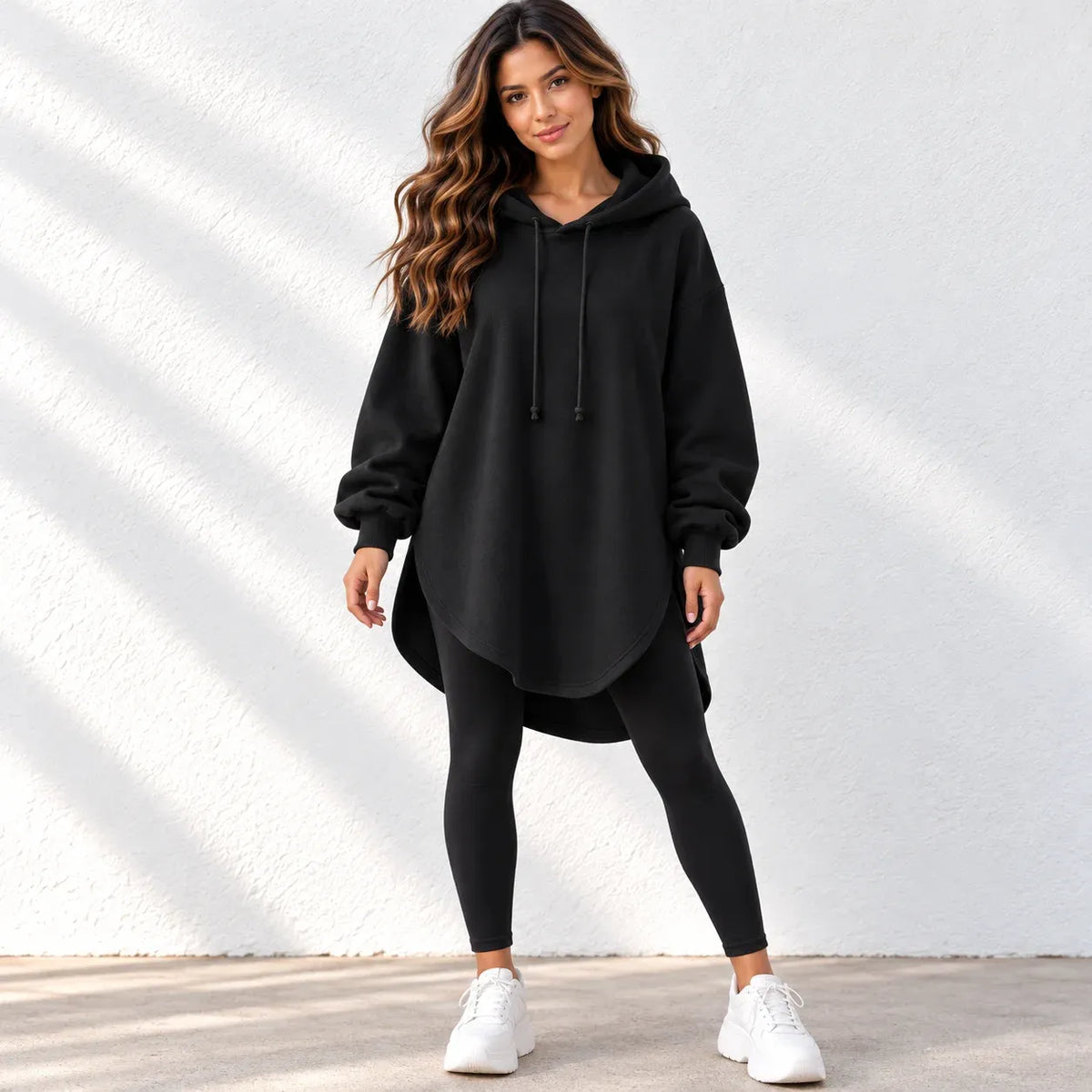 A woman modeling a black oversized pullover hoodie with drawstrings, paired with black leggings and white sneakers, standing against a white textured wall with soft diagonal shadows.