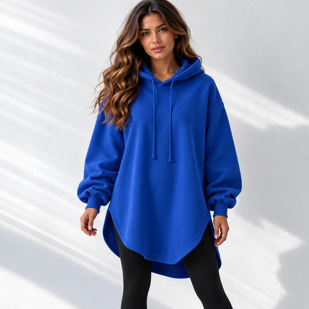 A woman wearing an oversized royal blue pullover hoodie with drawstrings and black leggings, standing against a white wall with soft diagonal shadows.